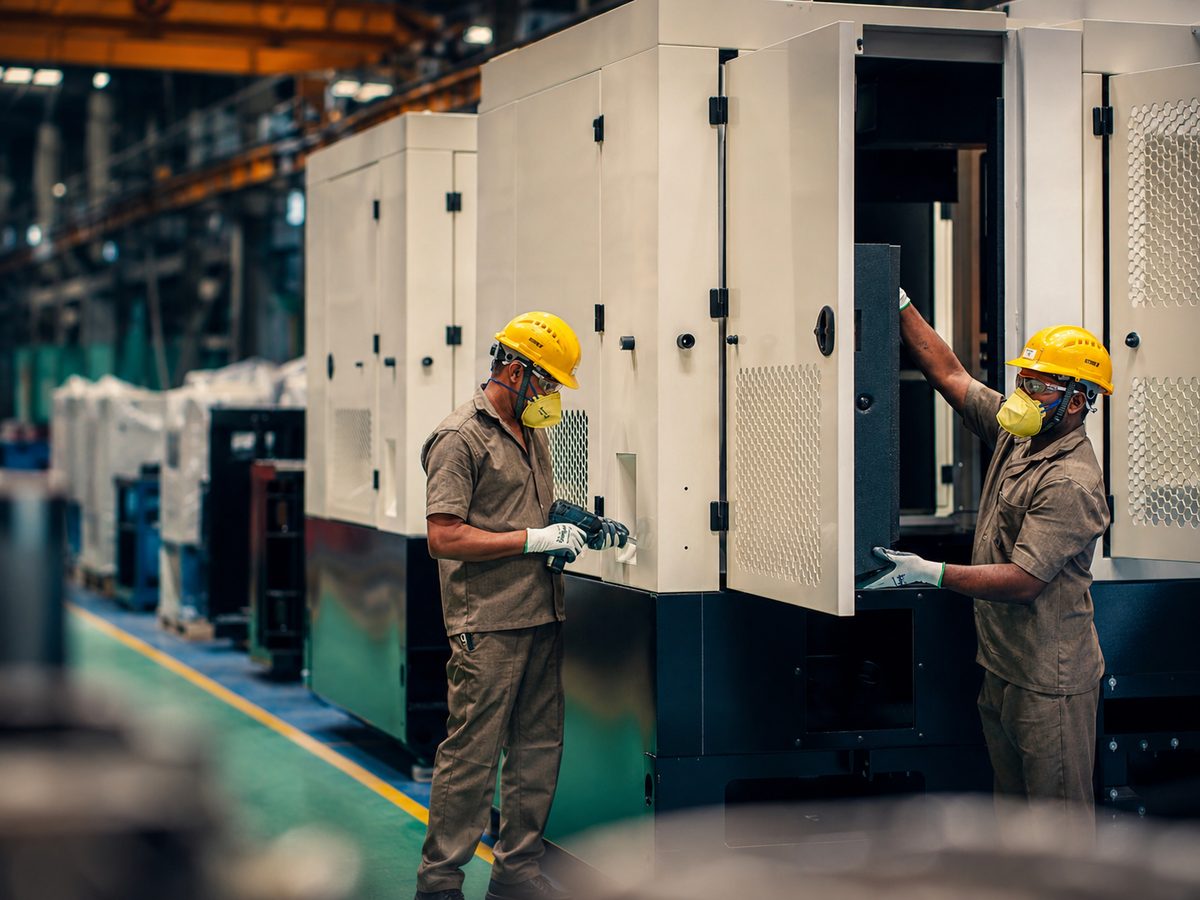 Two workers in hard hats and respirators fitting doors onto a large cream acoustic enclosure, with more enclosures receding down the bay